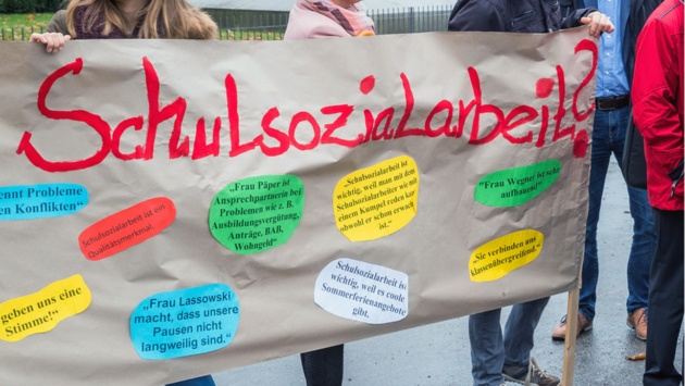 Teilnehmer einer Demonstration halten ein Plakat mit der Aufschrift "Schulsozialarbeit" hoch.