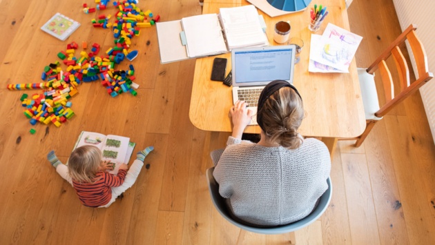 Ein zweijähriges Kind spielt im Wohnzimmer, während seine Mutter Zuhause im Homeoffice an einem Laptop arbeitet.